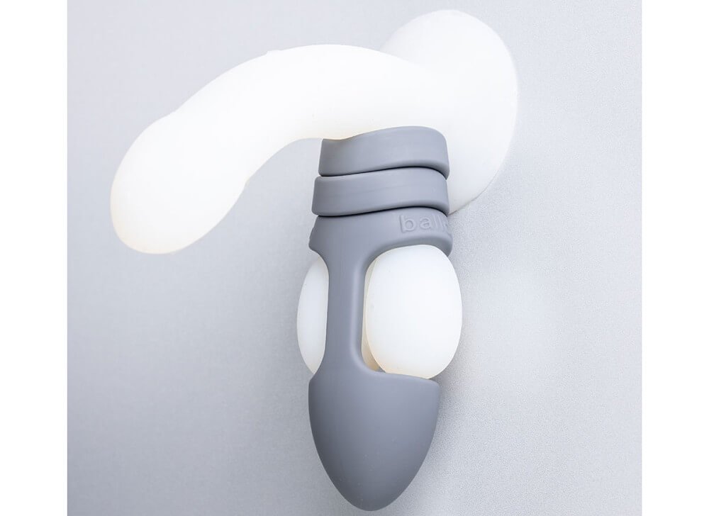 The Balldo in Steel Grey is shown being used on the testicles of a clear dildo. The image shows how the Balldo works - which is by wrapping around the testicles while simultaneously leaving space for the testicles to receive pleasure during penetration. | Kinkly Shop